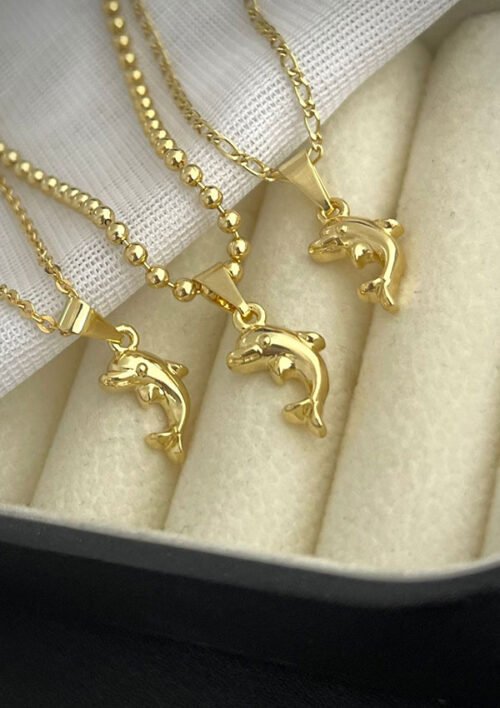 Tres collares de oro, cada uno con un colgante de delfín Dije, se exhiben sobre una tela de color crema. Estas exquisitas piezas son joyas en oro laminado de 18k.