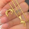 Una mano sostiene un collar de oro con el colgante de delfín Dije y un anillo de delfín dorado a juego, ambas exquisitas joyas en oro laminado de 18k. Las uñas de la persona están cuidadas con las puntas blancas.