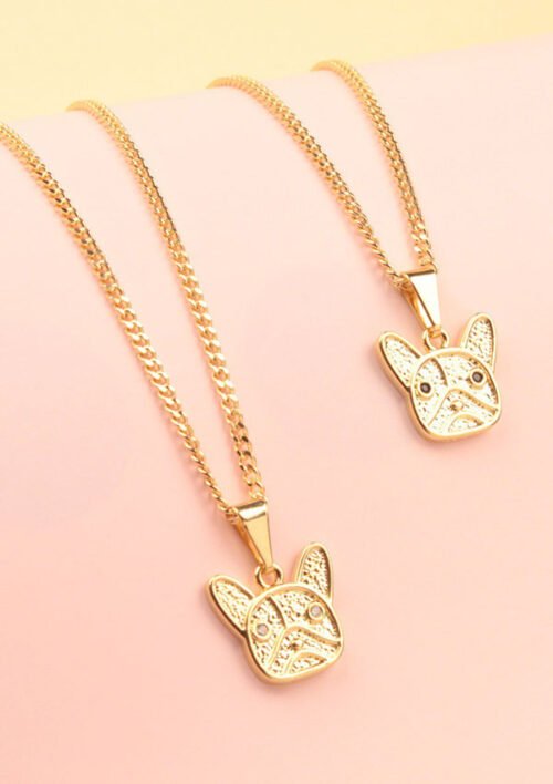 Dos cadenas de oro con dijes colgantes en forma de caras de perros, colocados sobre una superficie rosa, elaborados a partir de joyas en oro laminado de 18k.