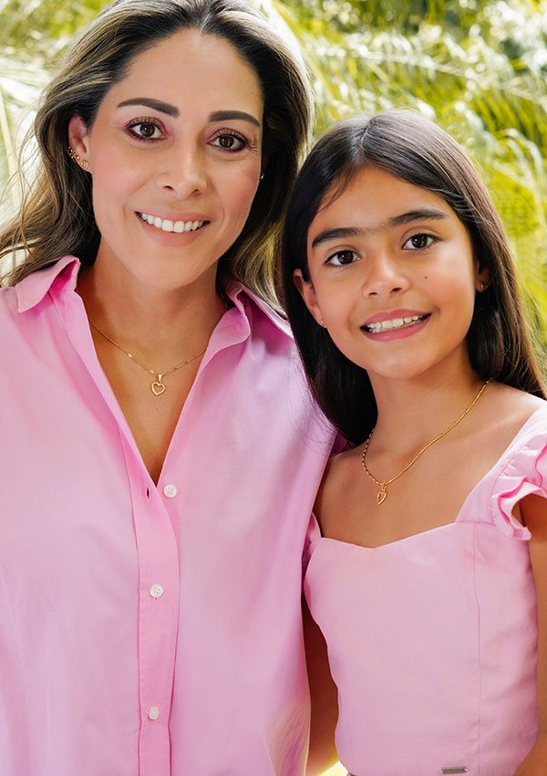 Una mujer y una niña están paradas una al lado de la otra, ambas sonrientes y con blusas rosas. Cada una tiene un collar de oro con un colgante en forma de corazón, elaborado con joyas de oro laminado de 18 quilates, conocido como el Dije Little Heart. Se ve follaje verde en el fondo.