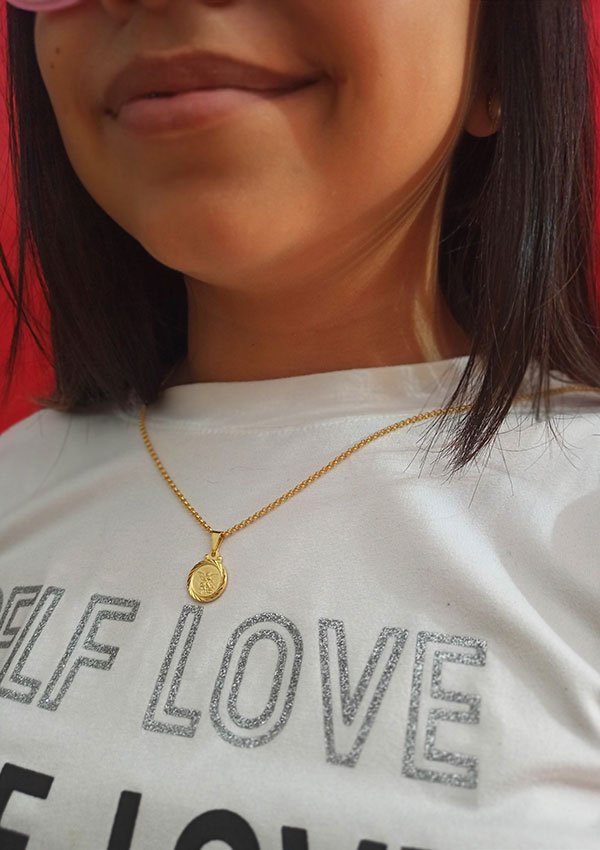 Una persona que viste una camisa blanca con un texto parcial de "AMOR PROPIO" y un collar de oro con un colgante circular, elaborado con joyas en oro laminado de 18k, con el Dije San Miguel Arcángel.