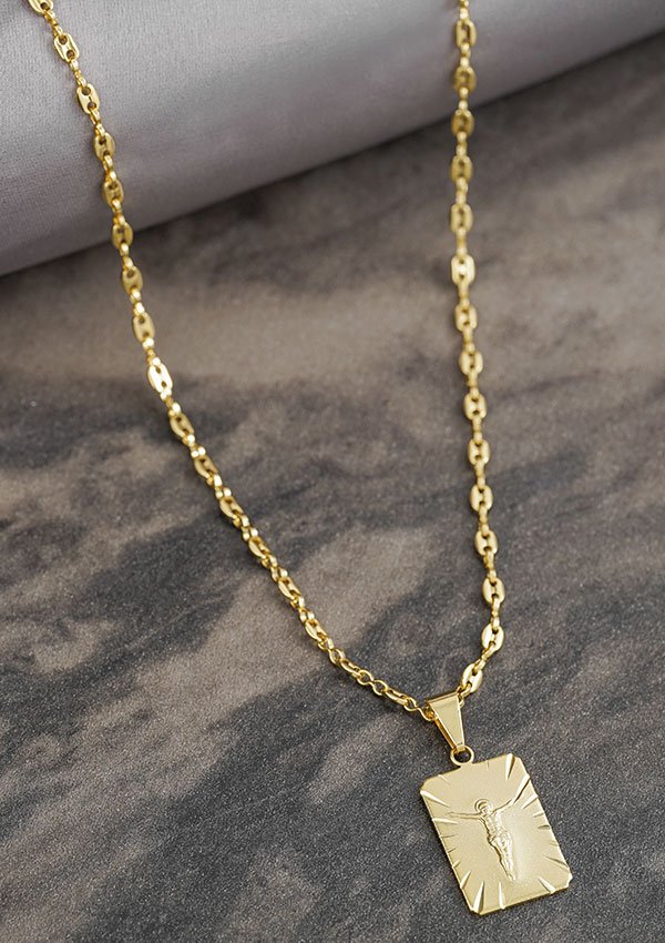 Collar colgante de oro con dije rectangular que presenta un diseño grabado de una figura. Esta exquisita pieza, de la colección Joyas en Oro Laminado 18K, se exhibe sobre una superficie de mármol oscuro.