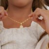 Un primer plano de un niño que sostiene un collar de oro con un colgante redondo adornado con diamantes mientras usa una blusa blanca texturizada muestra la elegancia de Joyas en Oro Laminado 18K.