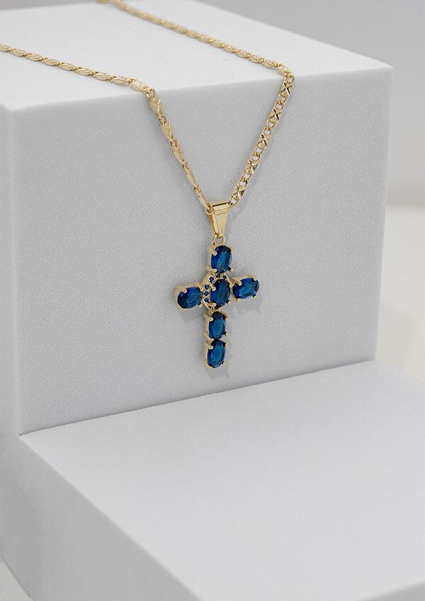 Un collar de oro con un colgante de cruz adornado con piedras preciosas azules, exhibido en un soporte de joyería blanco, ejemplifica la delicada artesanía de Joyas en Oro Laminado 18K.