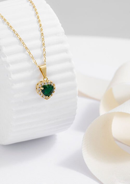 Un collar de oro con el colgante Dije Green Heart, adornado con una gema verde en forma de corazón rodeada de pequeñas piedras blancas, se exhibe sobre una superficie blanca texturizada. Una cinta pálida se enrolla a su lado, enfatizando la exquisita artesanía de las joyas en oro laminado de 18k.