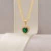 El "Dije Corazón Verde", un collar de cadena de oro que presenta un colgante verde en forma de corazón bordeado por pequeñas piedras blancas y exhibido en una cinta de color crema, ejemplifica la elegancia de las joyas en oro laminado de 18k.