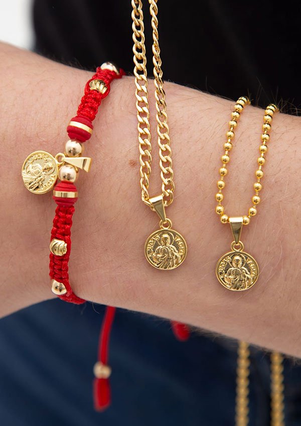 Primer plano de una muñeca adornada con dos pulseras de cadena de oro y una pulsera de cordón rojo, cada una con un pequeño colgante redondo de oro con un diseño grabado. Se trata de unas exquisitas joyas de "Dije San Judas Tadeo" en oro laminado de 18k, que añaden un toque de elegancia a cualquier look.