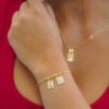 Persona que lleva una pulsera de oro con dos pequeños colgantes rectangulares y un collar a juego, cada uno con una imagen grabada. Joyas en Oro Laminado 18K añaden un toque elegante a este hermoso conjunto.