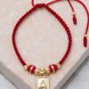 Pulsera trenzada roja que incluye un pequeño colgante rectangular de oro con una figura grabada, cuentas de oro y dos extremos de hilo rojo colgantes con detalles en oro. Esta impresionante pieza es parte de nuestra exquisita colección Joyas en Oro Laminado de 18K.