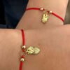 Dos muñecas llevan pulseras de hilo rojo con dijes de dragones de oro. Estas Joyas en Oro Laminado de 18K presentan pequeñas cuentas de oro y un adorno de dragón central.