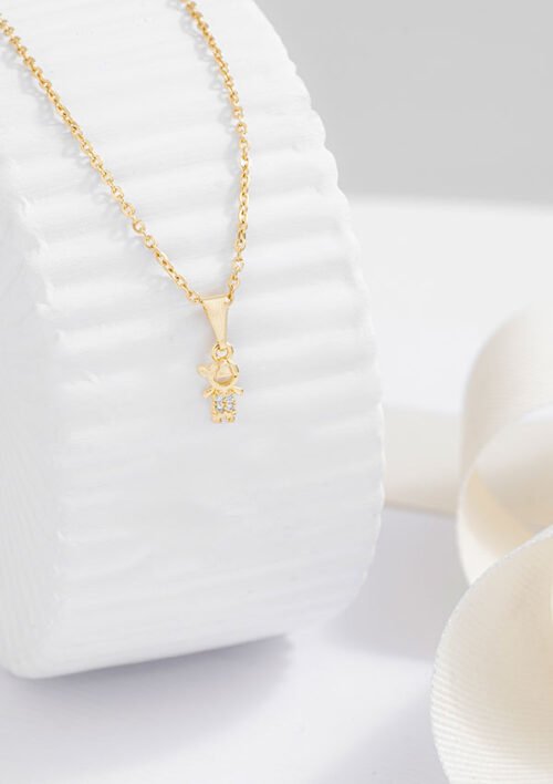 Un collar de cadena de oro con un pequeño colgante adornado con una diminuta piedra preciosa se exhibe elegantemente sobre una superficie cilíndrica con textura blanca, mostrando la exquisita artesanía de las joyas Dije Little Boy en oro laminado de 18k.