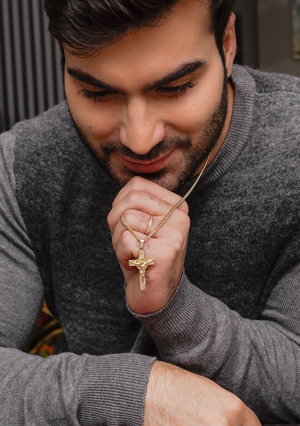 Un hombre con barba y vestido con un suéter gris sostiene un collar de oro con un colgante de cruz cerca de su rostro, mostrando el exquisito Dije Cruz San Benito joyas en oro laminado 18k.