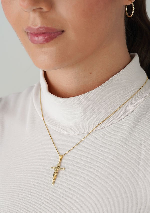 Primer plano de una persona que lleva un jersey de cuello alto blanco y un collar de crucifijo de oro, elaborado con Dije Madero Pequeño por Joyas en oro laminado de 18k. La persona muestra rasgos parcialmente visibles, incluidos los labios y un pendiente colgante.