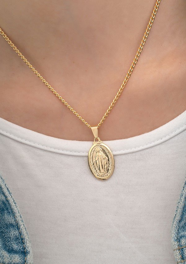 Primer plano del cuello de una persona que lleva un collar de cadena de oro en joyas en oro laminado 18k adornado con el "Dije Virgen Milagrosa Mediana" como colgante ovalado que presenta una figura religiosa, sobre una camisa blanca.