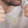 Una persona que lleva un suéter de punto sostiene un collar de oro con un colgante rectangular cerca de su cara, mostrando las joyas Dije El Señor es mi Pastor en oro laminado 18k.