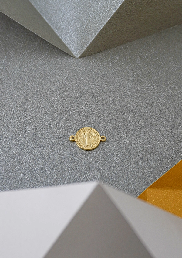 Una pequeña moneda de oro con dos presillas a cada lado reposa sobre una superficie gris texturizada, enmarcada por formas geométricas en el fondo, que recuerdan a Joyas en Oro Laminado de 18K.