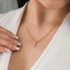 Primer plano de una persona que lleva un collar de oro con un colgante en forma de "D" y una parte superior blanca. La persona tiene las uñas perfectamente cuidadas y sostiene con delicadeza el collar, que es una de las impresionantes joyas en oro laminado de 18 quilates.