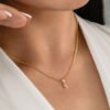 Primer plano del cuello de una mujer que lleva un collar de oro con un pequeño y elegante colgante en forma de letra 'S'. La mujer lleva una blusa blanca y se toca suavemente el cuello, mostrando sus exquisitas joyas en oro laminado de 18k.