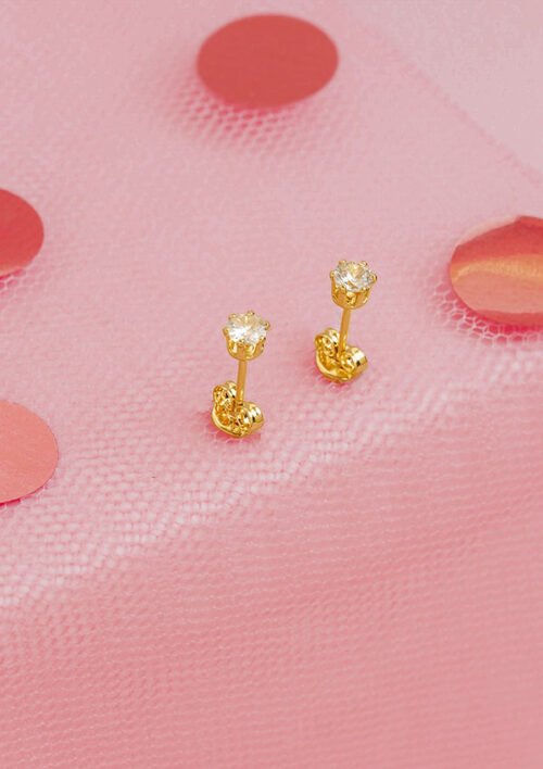 Un par de aretes de oro con piedras preciosas transparentes, presentados sobre una tela de malla rosa adornada con lunares rojos, que personifican la elegancia de las joyas en oro laminado de 18k.