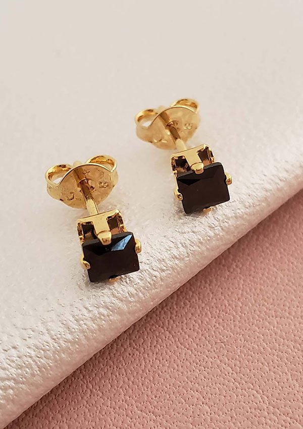 Los pendientes de oro con gemas cuadradas negras descansan elegantemente sobre una superficie suave y de color claro. Elaboradas con joyas de oro laminado de 18 quilates, estas piezas atemporales añaden un toque de sofisticación a cualquier conjunto.