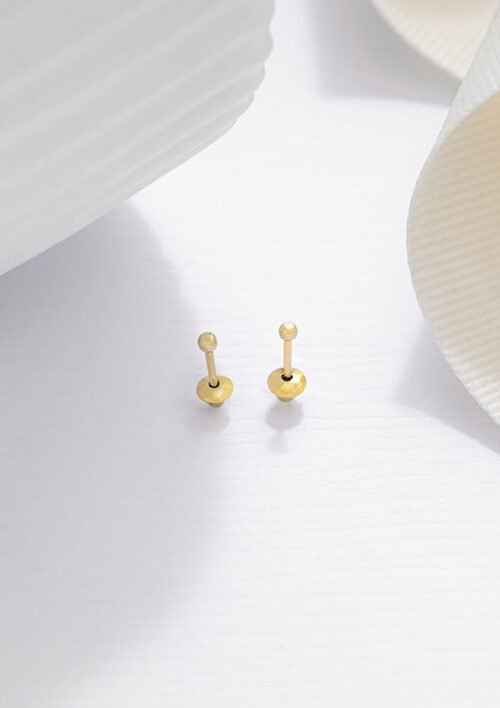 Dos impresionantes aretes Aretes Balin #2 de oro se disponen sobre una superficie blanca, acompañados de un objeto acanalado blanco y una delicada cinta al fondo. Estas exquisitas piezas de joyería, con oro laminado de 18 quilates, añaden un toque elegante a cualquier conjunto.