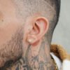 Primer plano de la oreja de una persona adornada con tres Aretes Balin #2 en oro laminado 18K y un corte de pelo afeitado, revelando tatuajes detallados en el costado de la cabeza y el cuello. Los intrincados diseños incluyen una rosa, entrelazada a la perfección con las elegantes joyas.