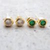 Dos pares de aretes tipo botón de Joyas en Oro Laminado de 18K, uno engastado con piedras preciosas redondas blancas y el otro engastado con piedras preciosas redondas verdes, colocadas sobre una superficie gris.