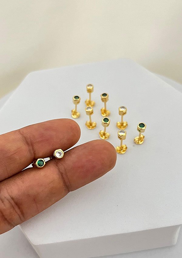 Una mano sostiene dos pequeñas piezas de joyería, una con una piedra verde y otra con una piedra blanca. En el fondo se muestran varias Joyas en Oro Laminado 18K similares sobre una superficie blanca.