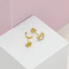 Un par de aretes de oro con pequeñas piedras transparentes se muestran sobre un fondo geométrico blanco y rosa, mostrando la elegancia de Joyas en Oro Laminado 18K.