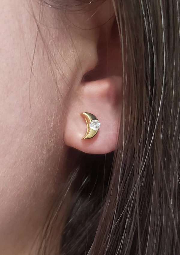 Primer plano de una oreja adornada con un pendiente de media luna de oro, engastado con un pequeño diamante, de la colección Joyas en Oro Laminado 18K.