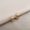 Dos pequeños aretes en forma de media luna de oro con piedras incrustadas, parte de nuestra colección Joyas en Oro Laminado 18K, están colocados sobre una superficie gris claro texturizada.