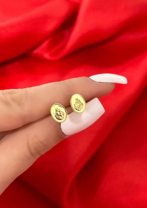 Primer plano de una mano que sostiene un par de pequeños aretes de oro con un diseño detallado, sobre un fondo de tela roja. Joyas en Oro Laminado 18K.