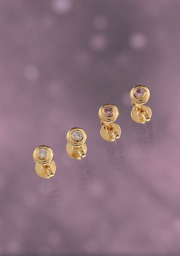 El conjunto de cuatro Aretes Circon Caja Redonda de cristal de 4 mm, con piedras preciosas redondas, elegantemente dispuestas en línea sobre una superficie reflectante de color lavanda, muestra la sofisticación de las joyas de oro laminado de 18 quilates.