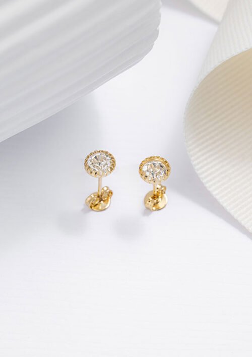 Los Aretes Topo Circon de 6 mm están elegantemente colocados sobre una superficie blanca con una cinta cerca, mostrando la belleza atemporal de las joyas elaboradas en Oro laminado de 18 quilates.