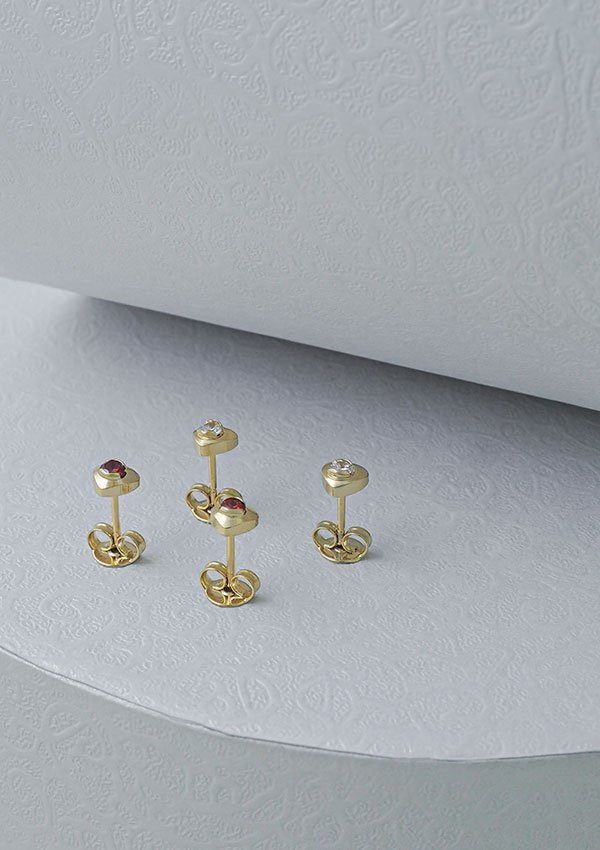 Tres tachuelas de oro en forma de corazón con detalles de piedras preciosas colocadas sobre una superficie blanca texturizada muestran la elegancia de las joyas en Oro laminado de 18 quilates.