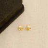 Un par de aretes en forma de flor de oro con piedras preciosas transparentes se exhiben junto a una caja de joyería negra sobre una superficie de tela beige, mostrando exquisitas Joyas en Oro Laminado de 18K.