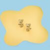 Dos Aretes Osito Teddy, aretes de oro con forma de osito elaborados con Oro laminado 18K, se muestran sobre una superficie de color amarillo claro sobre un fondo azul claro.