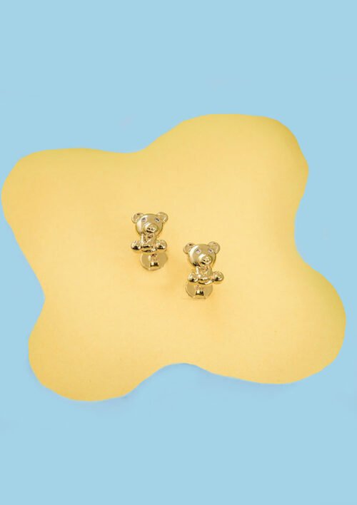 Dos Aretes Osito Teddy, aretes de oro con forma de osito elaborados con Oro laminado 18K, se muestran sobre una superficie de color amarillo claro sobre un fondo azul claro.