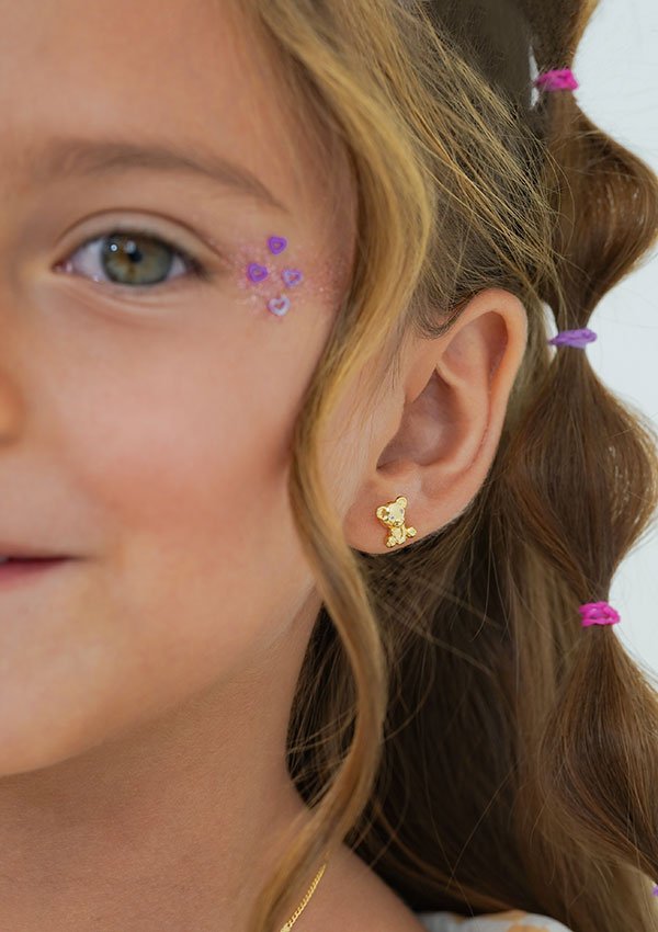 Primer plano del rostro de un niño, centrándose en su oreja adornada con un pendiente Aretes Osito Teddy de oro laminado de 18K con forma de osito. La niña tiene el pelo castaño claro decorado con pequeñas trenzas atadas con bandas de colores y lleva pegatinas en la cara con corazones de color púrpura.