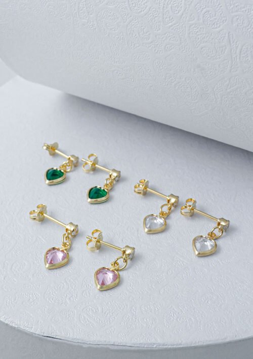 Tres pares de Aretes Corazón Circonias 1.7 Cms en oro, con colgantes en forma de corazón con gemas verdes, transparentes y rosas, se colocan sobre una superficie blanca texturizada.