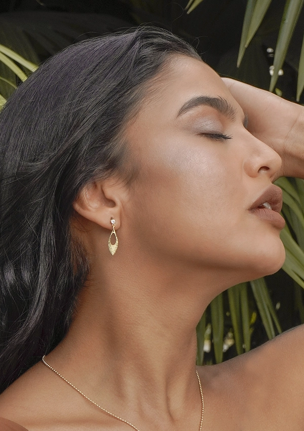 Una persona con cabello largo y oscuro lleva un collar de oro y un pendiente colgante, etiquetado Joyas en Oro Laminado 18K, con los ojos cerrados y la cabeza inclinada hacia atrás en medio del follaje verde.