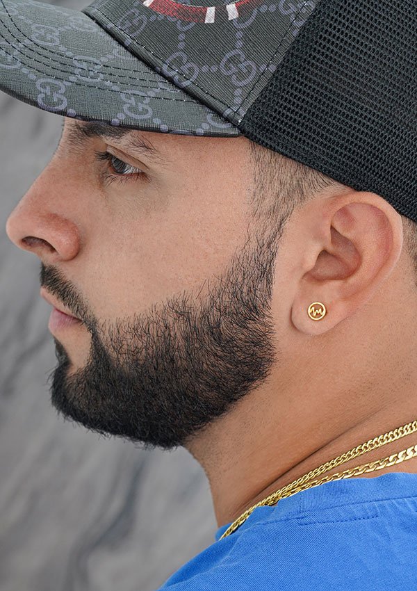 Perfil lateral de un hombre con gorra gris y negra, arete dorado, collar y camisa azul, con barba corta y bigote. Sus joyas Aretes Wave oro laminado 18K añaden un toque elegante a su look.