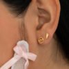 Primer plano de la oreja de una persona adornada con dos pequeños aretes de oro: uno redondo con diseño de ondas, conocido como Aretes Wave, y otro con forma de huella. Cerca de la oreja se ve una cinta de encaje blanco atada en un lazo, resaltando las delicadas joyas realizadas en Oro laminado 18K.