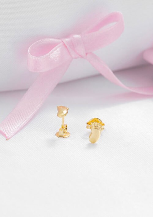 Dos pequeños "Aretes Baby Foot" dorados con detalles en rosa, elaborados en Oro laminado 18K, colocados sobre una superficie blanca, con una cinta rosa claro atada en un lazo en el fondo.