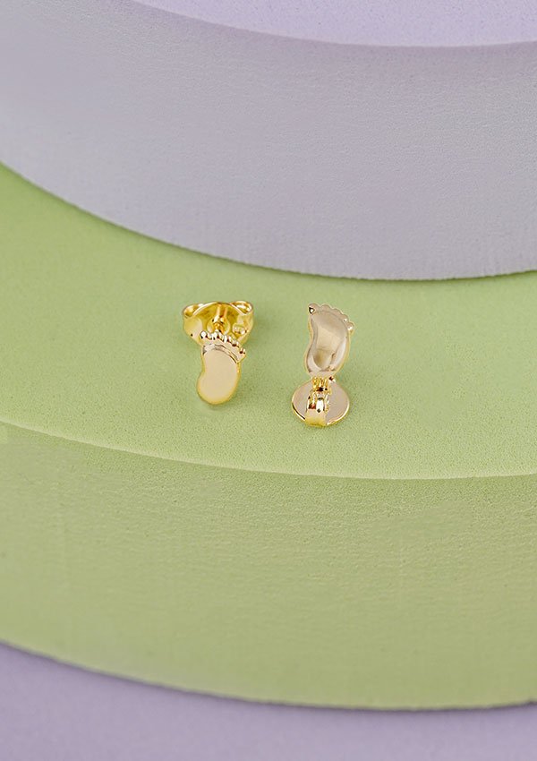 Dos Aretes Baby Foot, pendientes de oro con forma de pies de bebé y elaborados en Oro laminado de 18K, colocados sobre un fondo texturizado de color verde y morado.