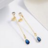 Aretes L Blue, con aretes colgantes de oro adornados con piedras preciosas azules en forma de lágrima elaboradas en Oro Laminado de 18 quilates, elegantemente colocadas sobre una superficie blanca cerca de una cinta blanca. Estas joyas capturan elegantemente la sofisticación y la belleza atemporal.