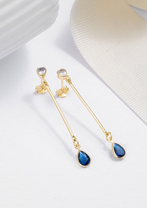 Aretes L Blue, con aretes colgantes de oro adornados con piedras preciosas azules en forma de lágrima elaboradas en Oro Laminado de 18 quilates, elegantemente colocadas sobre una superficie blanca cerca de una cinta blanca. Estas joyas capturan elegantemente la sofisticación y la belleza atemporal.