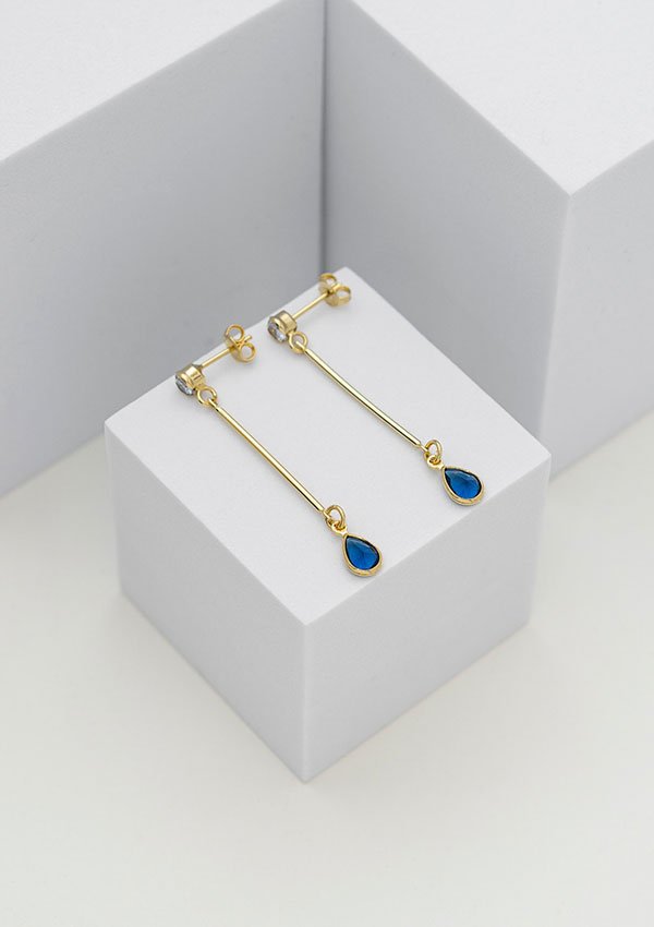 Un par de aretes colgantes de oro laminado de 18 quilates Aretes L Blue con piedras azules en forma de lágrima, elegantemente expuestas sobre bloques geométricos blancos.