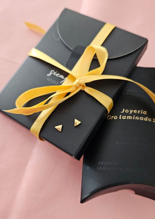 Cajas de regalo negras con cintas amarillas, una de ellas contiene un par de aretes triangulares pequeños de Joyas en Oro Laminado de 18K. Las cajas están colocadas sobre una superficie rosa.