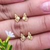 Primer plano de una mano con uñas cuidadas sosteniendo dos pares de aretes de mariposa de oro con piedras centrales verdes y transparentes, elaborados con Joyas en Oro Laminado de 18K, colocados en medio de follaje verde y flores blancas.